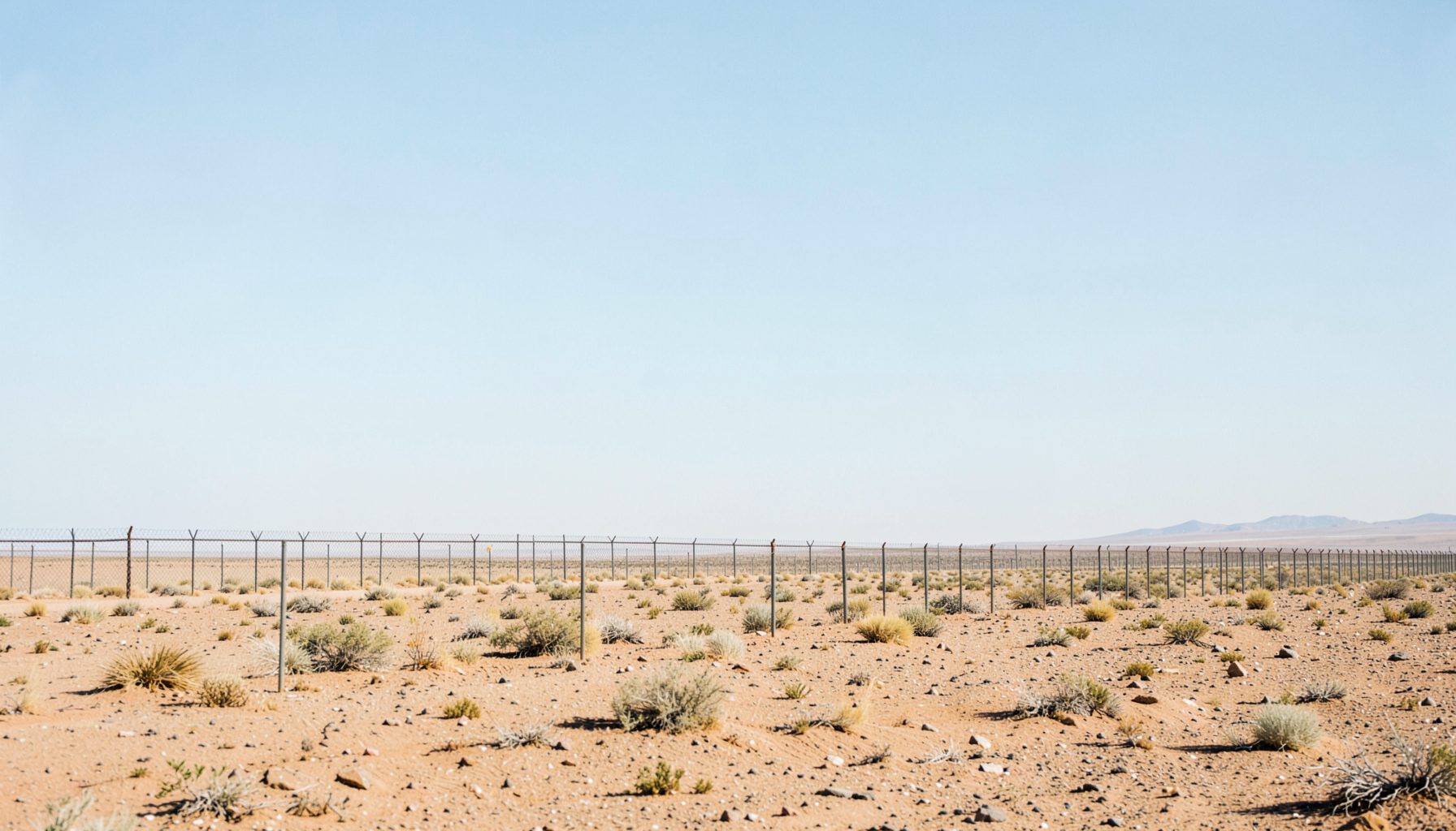 A panoramic desert landscape with a border fence stretching into the distance under a wide, pale-blue sky, suggesting isolation and vastness.
