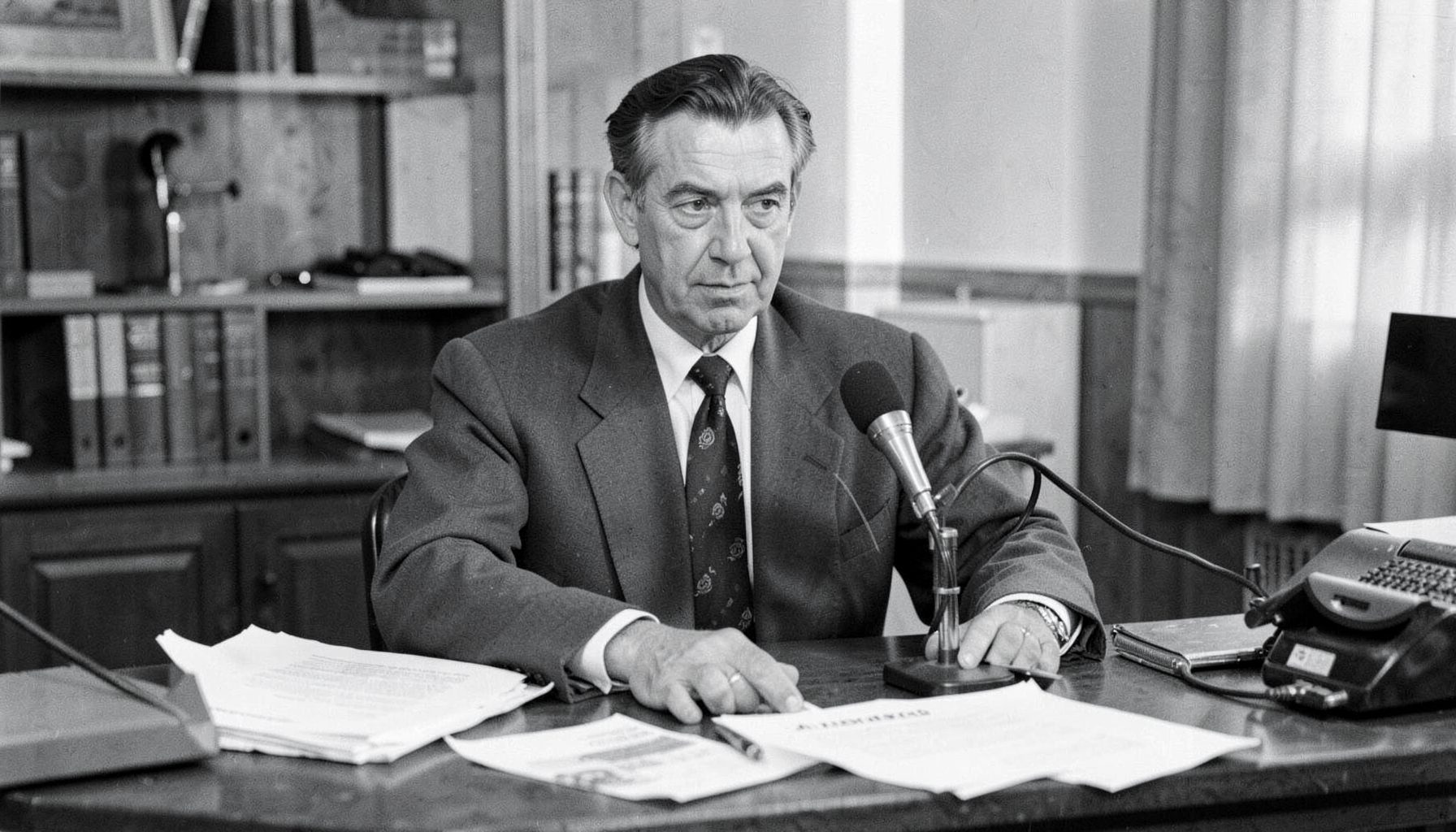 A reporter at a desk with papers and a microphone, black and white image conveying the atmosphere of mid-century Israeli journalism